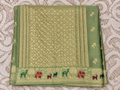 A folded sage green silk saree with intricate gold zari  Banarasi Brocade Saree , featuring a decorative border with colorful elephant motifs in red, green, and blue, laid flat on a textured beige patterned fabric.