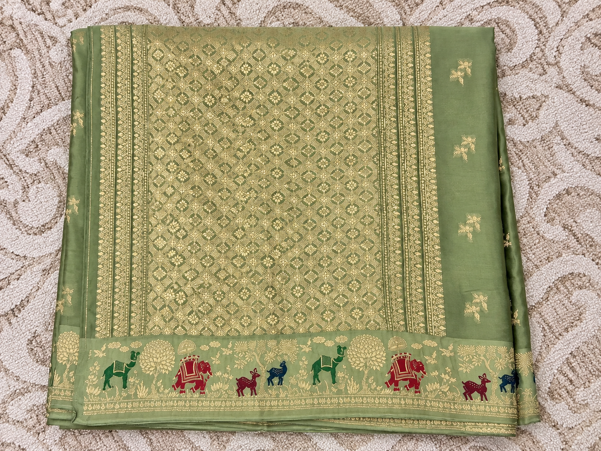 A folded sage green silk saree with intricate gold zari  Banarasi Brocade Saree , featuring a decorative border with colorful elephant motifs in red, green, and blue, laid flat on a textured beige patterned fabric.
