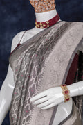 A close-up of a draped saree on a white mannequin, featuring a taupe-gray body with intricate metallic zari motifs and a contrasting deep maroon blouse. The mannequin is styled with a matching maroon beaded necklace and a gold bracelet with red stones.